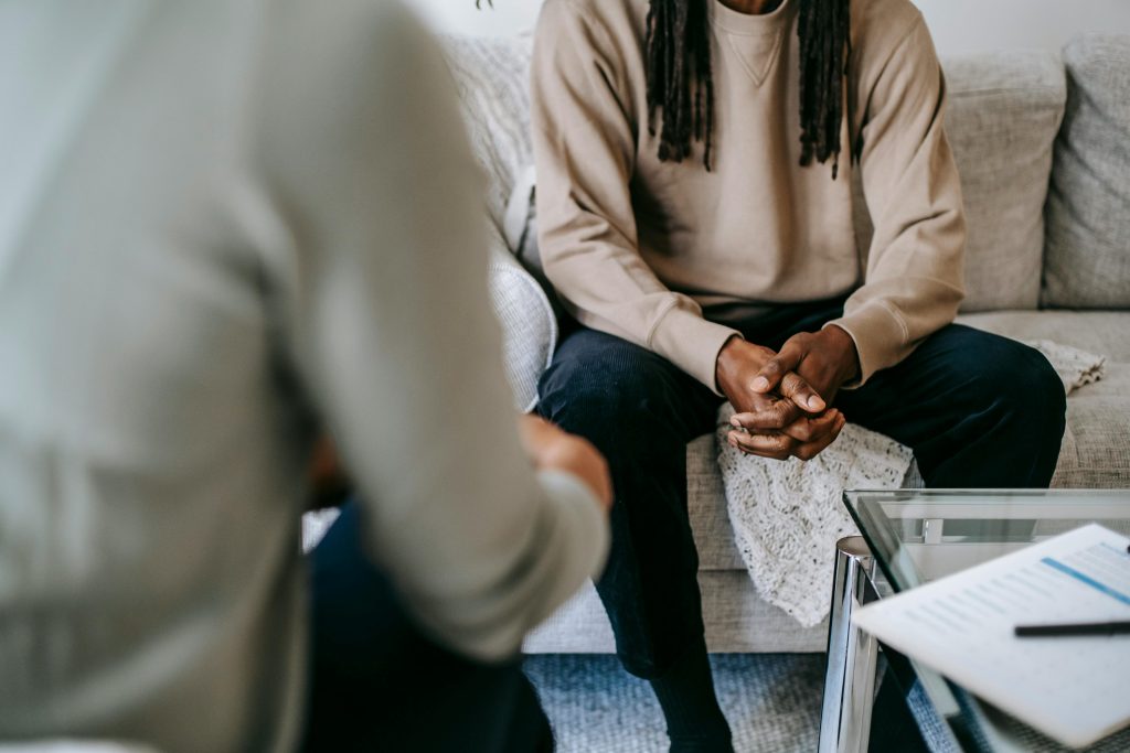 pexels photo 5699434 5699434 Unrecognizable African American male patient with clasped hands sitting on sofa near blurred anonymous therapist during visit in psychotherapy office