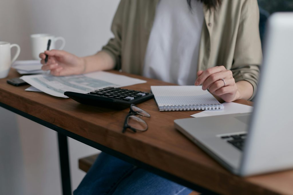 pexels photo 6963857 6963857 A woman manages finances at home, using a laptop and calculator on a wooden desk.